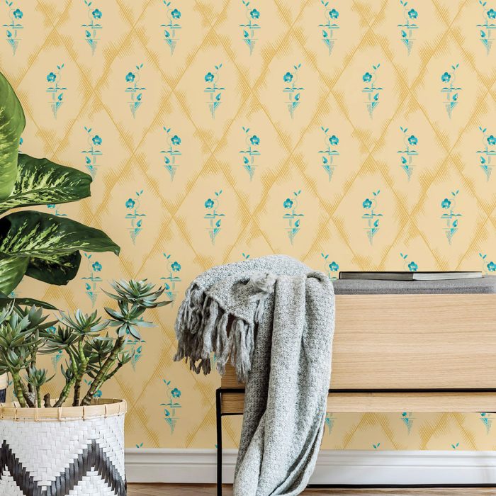 A Deco Diamond bench with a gray blanket sits before blue floral vintage wallpaper, flanked by a potted plant and leafy greenery for a charming set design.