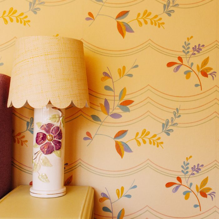 The Fiesta ceramic table lamp, featuring a floral design and woven shade, stands before vintage wallpaper adorned with colorful leaves and scalloped lines.