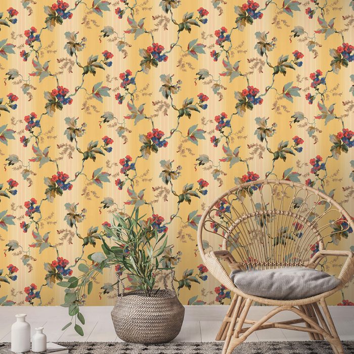 The Trentina wicker chair and a potted plant rest on a rug before vintage yellow floral wallpaper, creating a charming set design.