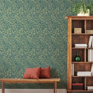 Against a green wall featuring the "Willow Boughs in Summer" motif, a wooden bookshelf holds books and a plant. In front, a bench with two red cushions enhances the cozy nook's charm.
